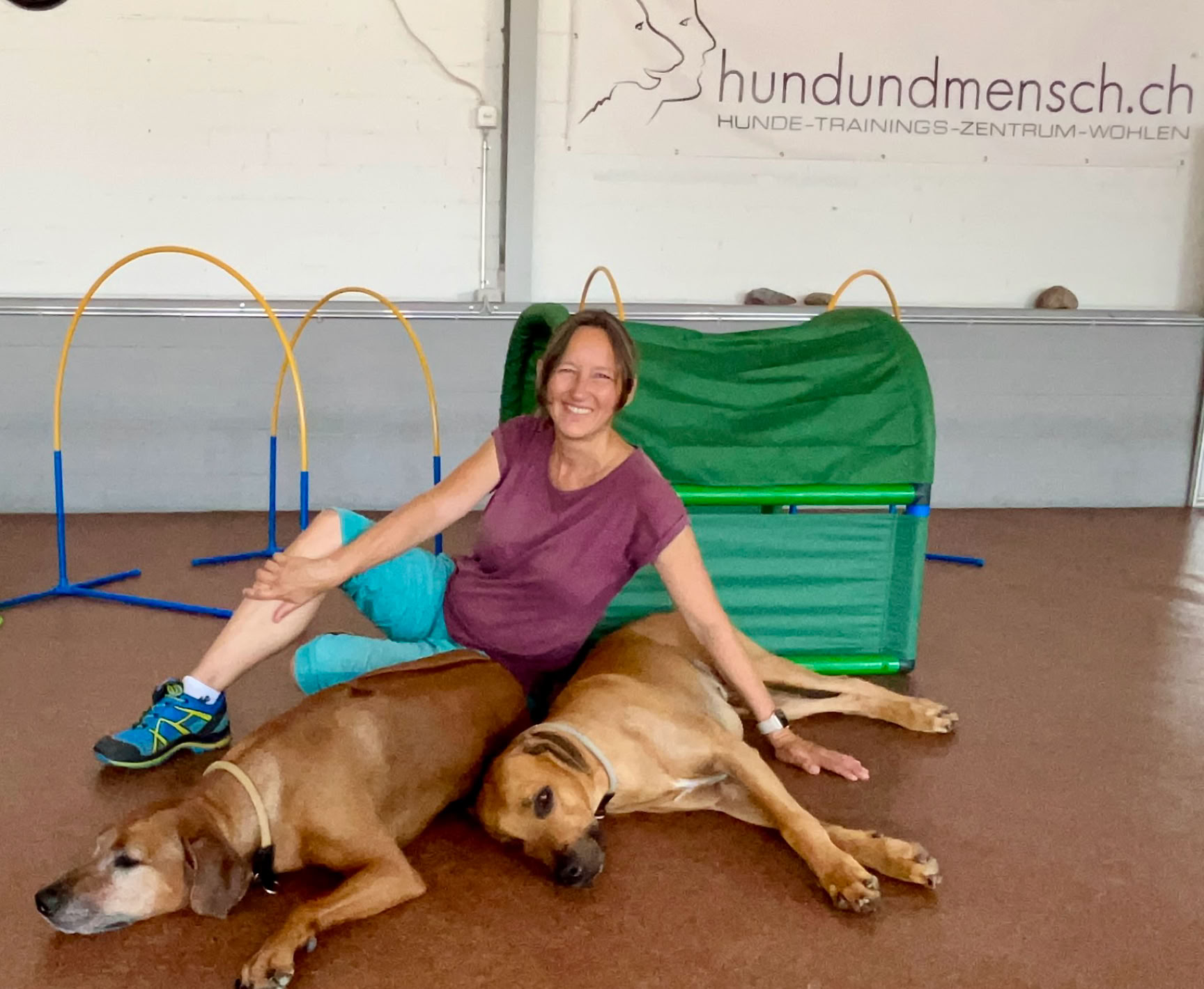 Erika sitzt auf dem Boden neben ihren beiden Rhosian Ridgeback Hunden Ema und Winston, die auf dem Boden liegen. Im Hintergrund sind ein grünes Hindernis und blaue Stangen zu sehen.