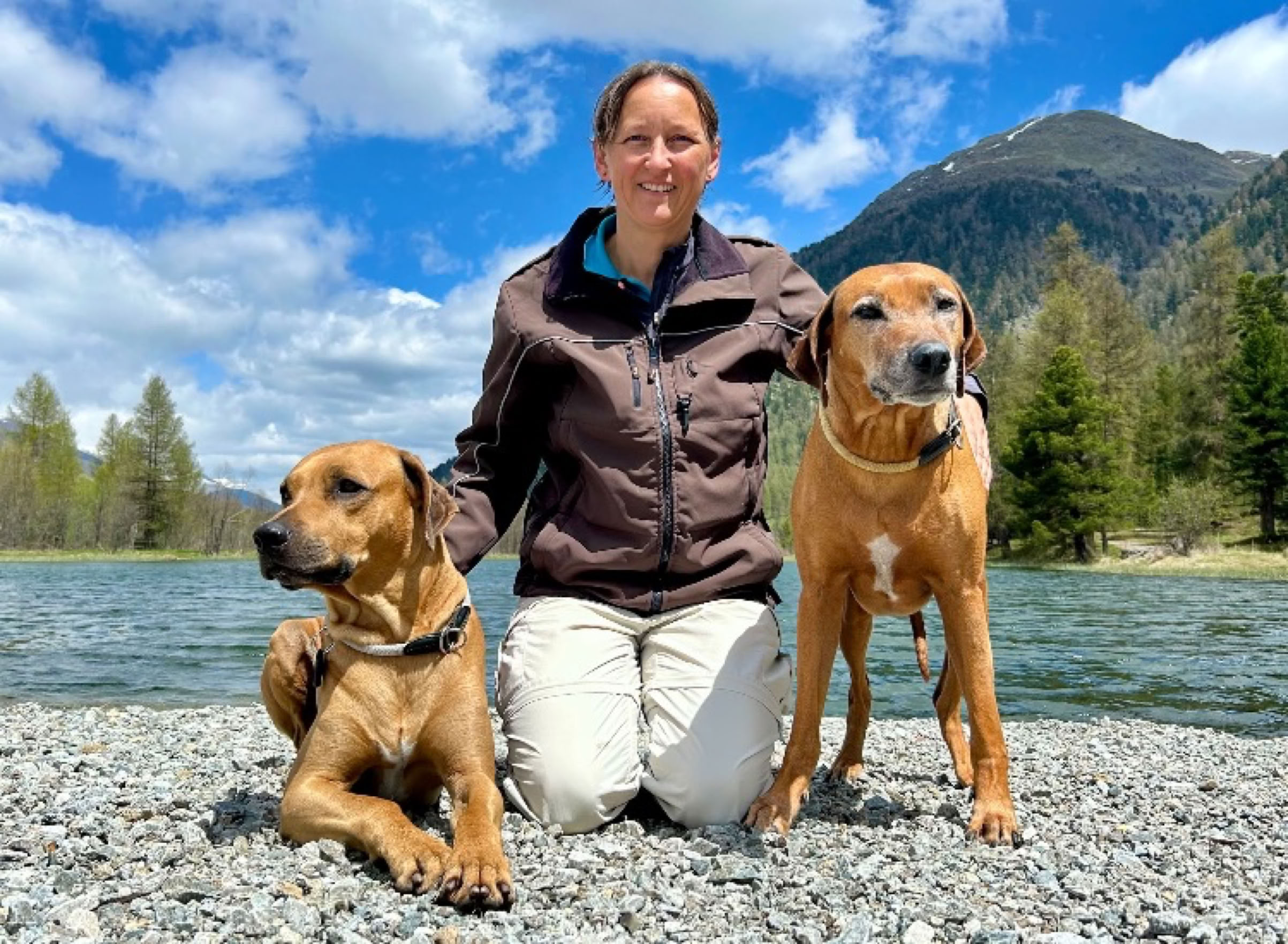 Erika Tauscher kniet auf einem Kiesstrand zwischen ihren zwei braunen Rhodesian Ridgeback, im Hintergrund ein See, Bäume und ein Berg unter blauem Himmel mit Wolken.