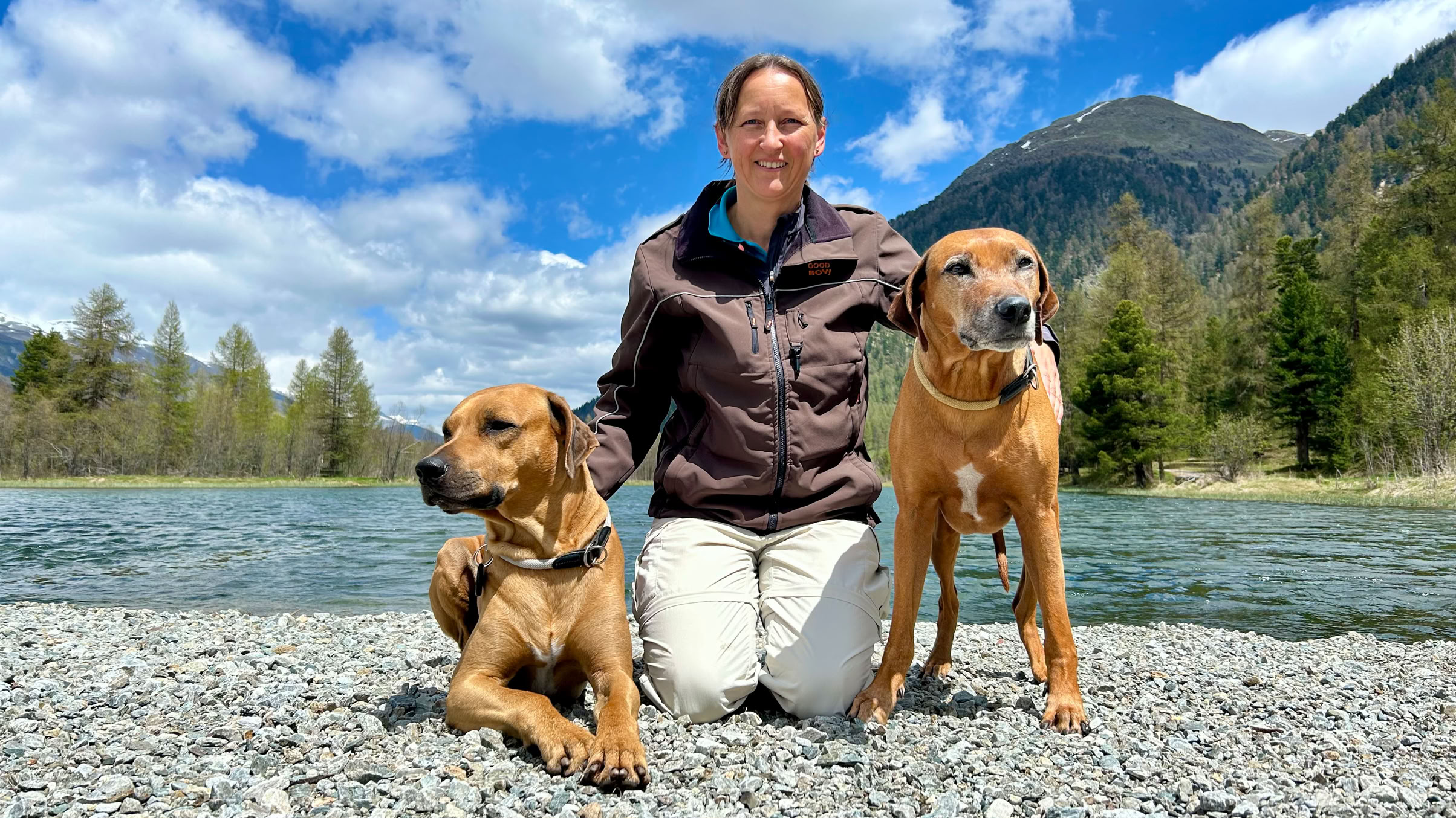 Erika kniet auf einem Kiesstrand zwischen ihren beiden Rhodesian Ridgeback Hunden Winston und Ema, im Hintergrund sind ein Fluss, Bäume und Berge unter einem blauen Himmel mit Wolken zu sehen.